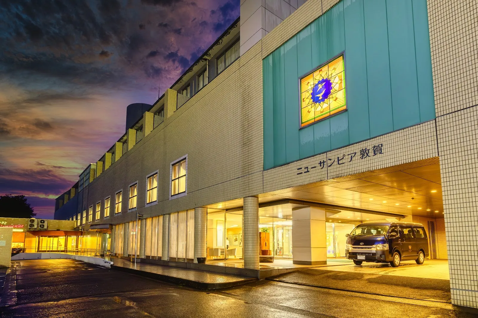 夕暮れの敦賀ニューサンピアの明るく照らされた正面。ガラス張りの入口と看板が駐車場越しに見えます。 — New Sunpia Tsuruga Ice Arena