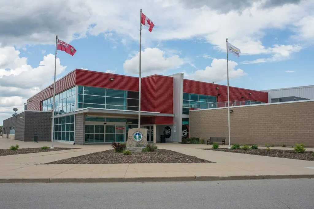 The glass and red-panel entrance with three flagpoles in front of the brick arena block. — New Tecumseth Recreation Centre