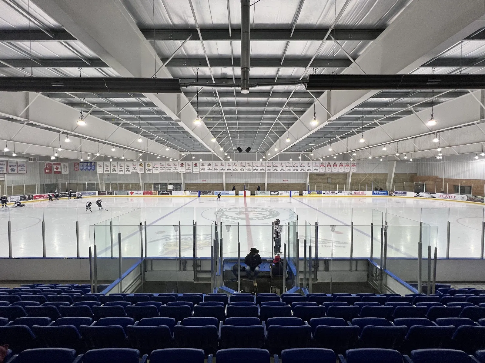 A full ice pad seen from rows of blue seats, with the glass and a second pad visible beyond under a white steel roof. — New Tecumseth Recreation Centre