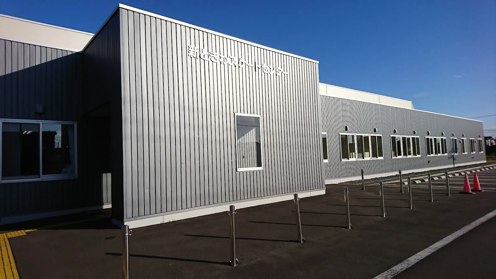 晴れた日の新ときわスケートセンターの、灰色の金属張りの建物。妻面に名称の看板があり、手前に黒い舗装の駐車場が広がっています。 — New Tokiwa Skate Center