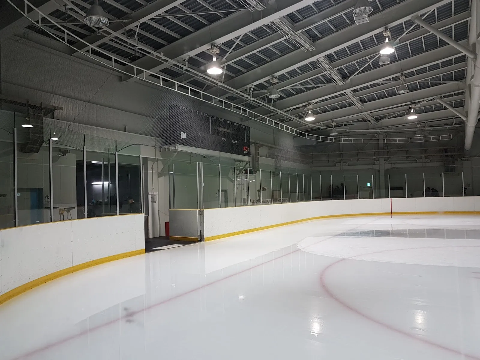 鉄骨トラスの屋根の下にある新ときわスケートセンターの空の氷面。黄色い縁のフェンスと、氷上のブルーラインとフェイスオフサークルが見えます。 — New Tokiwa Skate Center