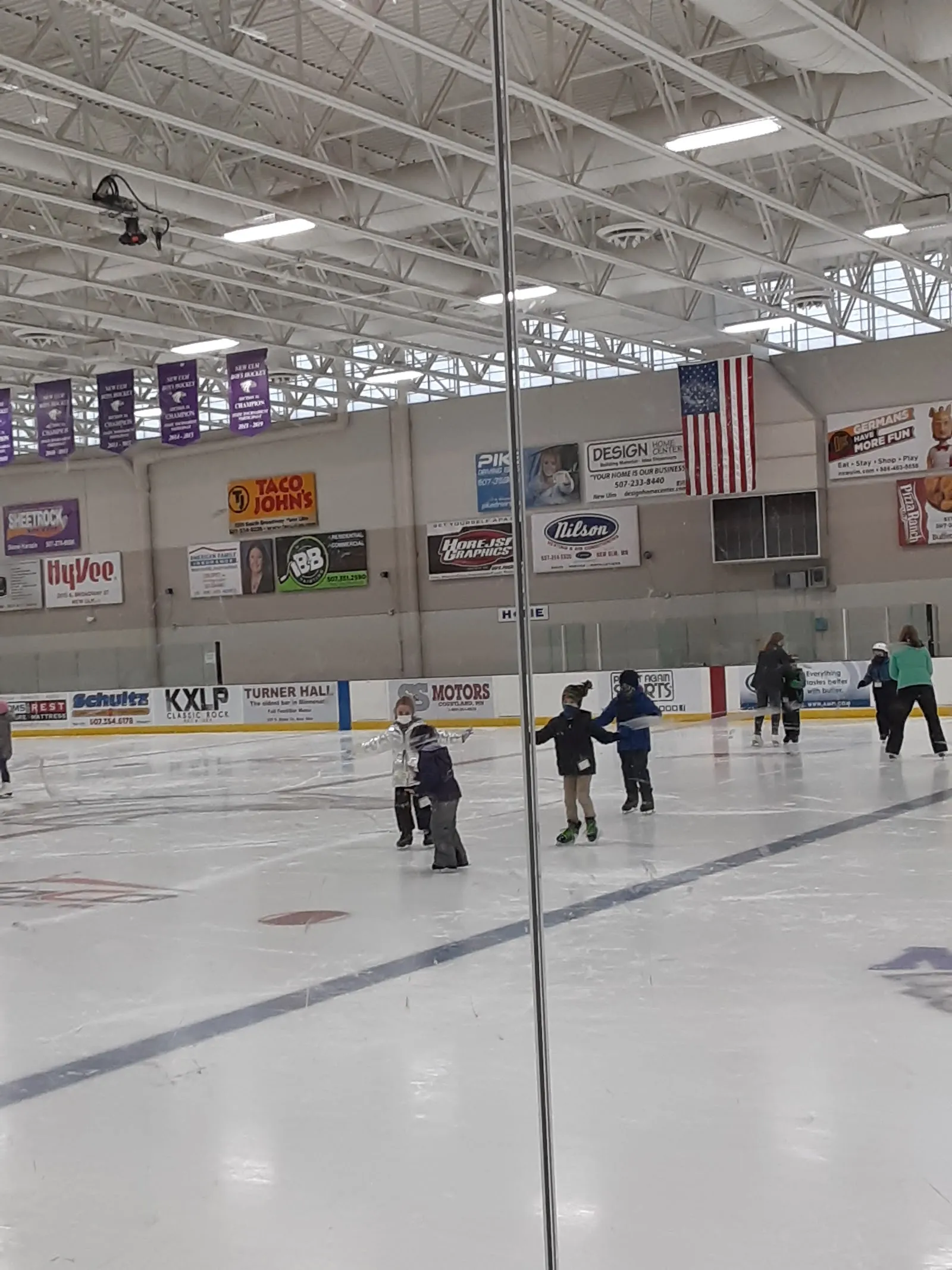 A game of skaters spread along the far side of the ice seen through a glass panel, sponsor boards and an American flag along the end wall under a long ceiling. — New Ulm Civic Center