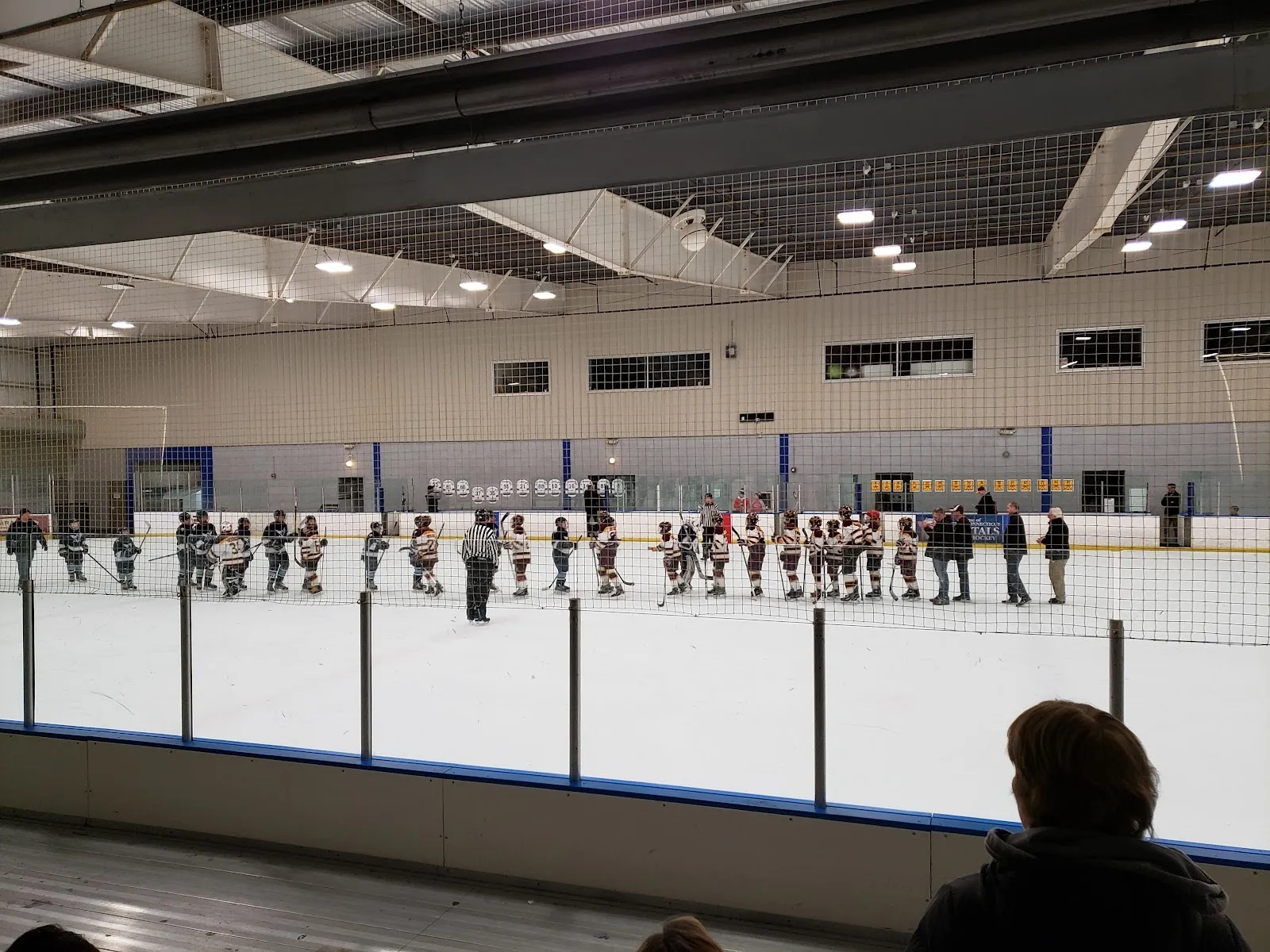 Players lined up along the blue line far across the sheet, seen over the glass under a pale ceiling with netting along the end wall. — Newington Arena