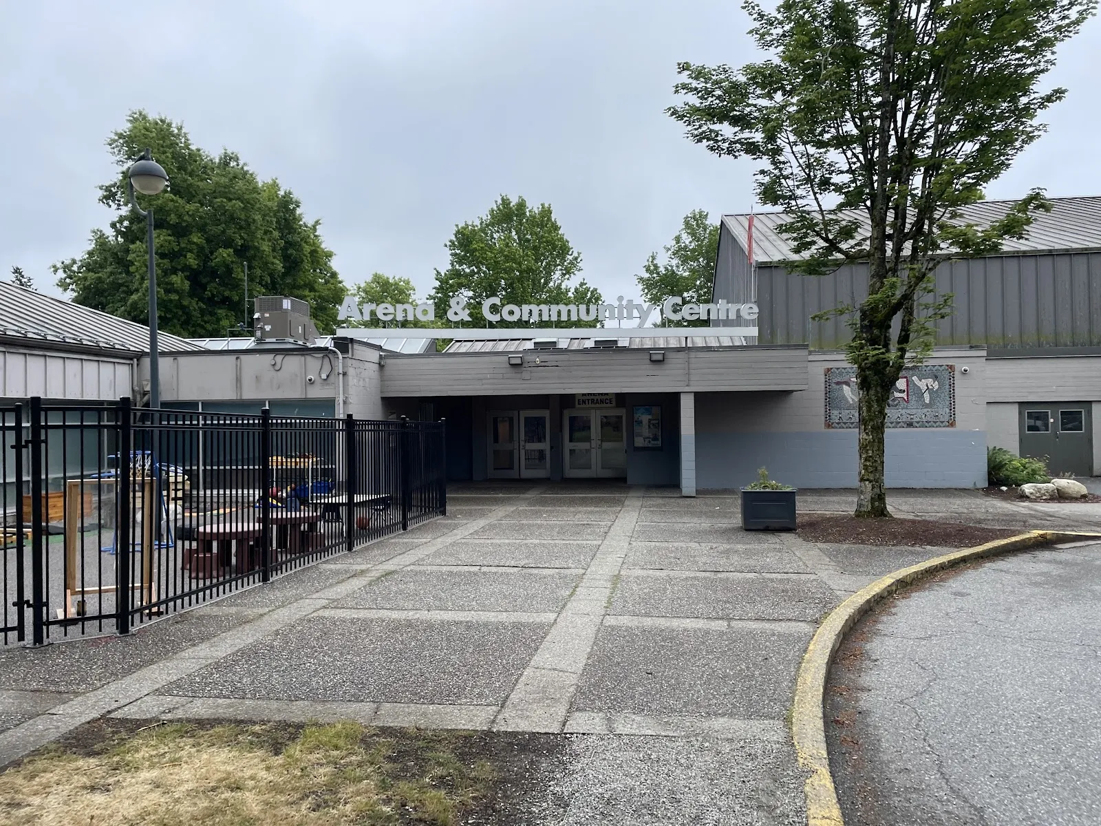 Front entrance walkway with a large "Arena & Community Centre" sign above the doors, a mosaic mural on the wall beside it, and a fenced playground area to the left. — Newton Arena