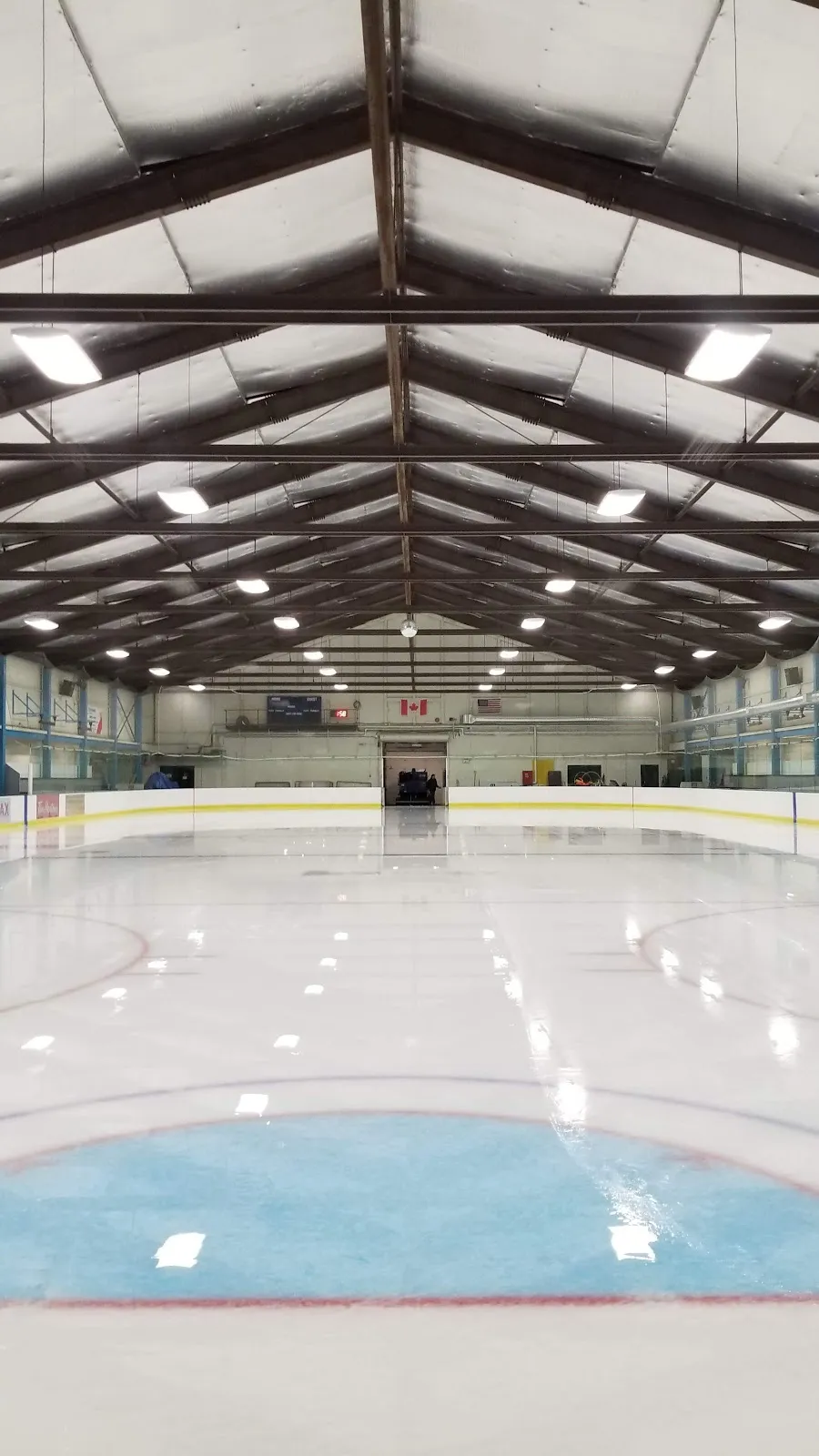 Empty ice sheet from ice level toward the far end, under a peaked steel-truss roof, with Canadian and U.S. flags on the end wall. — Newton Arena
