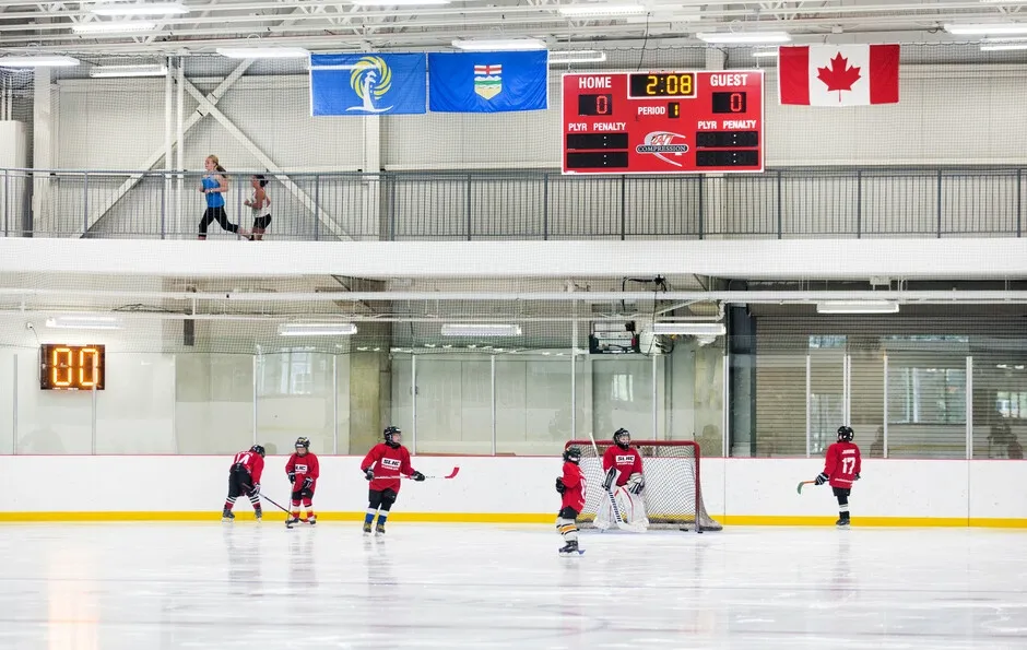 Young players in red around a net on the ice, a scoreboard and Alberta and Canadian flags hung above the glass behind them. — NexSource Centre