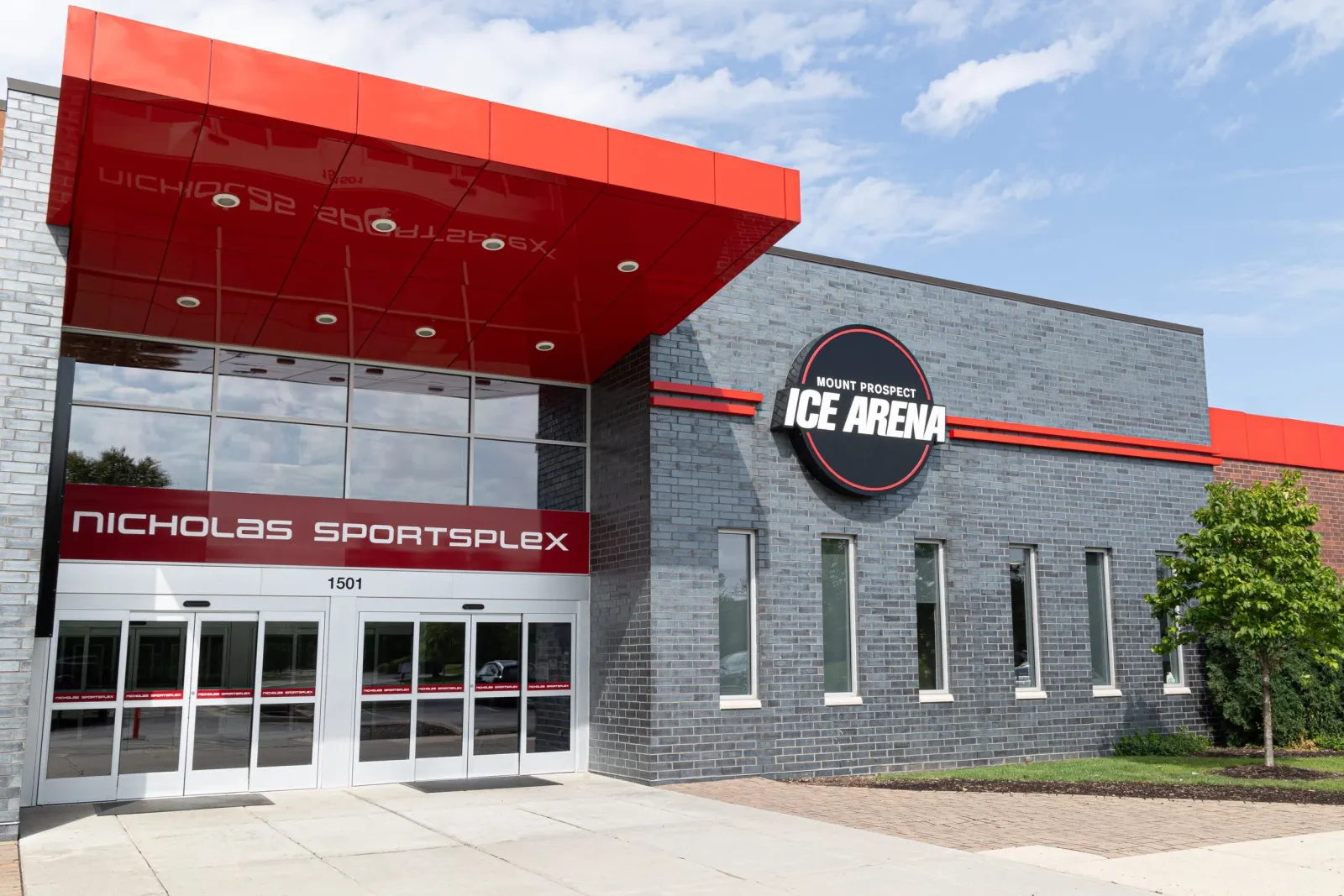 The main entrance on a clear day: a red cantilevered canopy over glass doors marked "NICHOLAS SPORTSPLEX" and the street number 1501, with the round "Mount Prospect Ice Arena" sign on the grey brick wall beside it. — Nicholas Sportsplex (Mount Prospect)