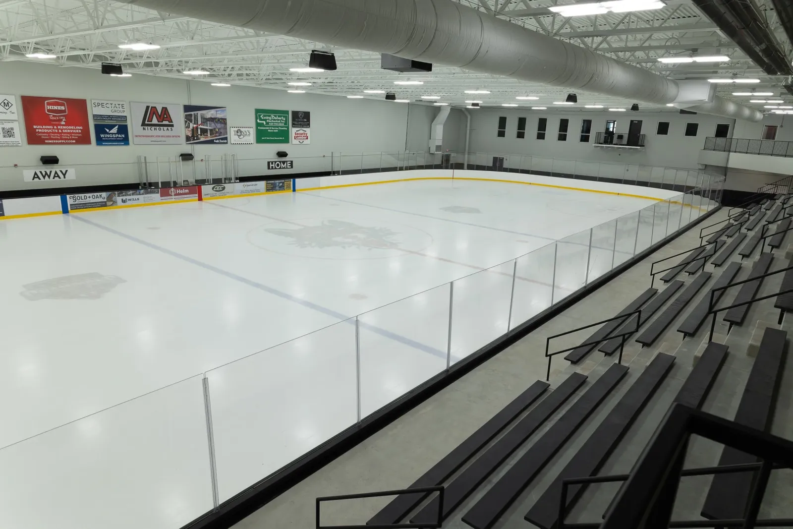 An empty sheet seen from the bleachers, with the centre-circle crest, HOME and AWAY benches, glass-topped boards, sponsor banners including the Nicholas construction banner, and a spectator mezzanine along the far wall. — Nicholas Sportsplex (Mount Prospect)