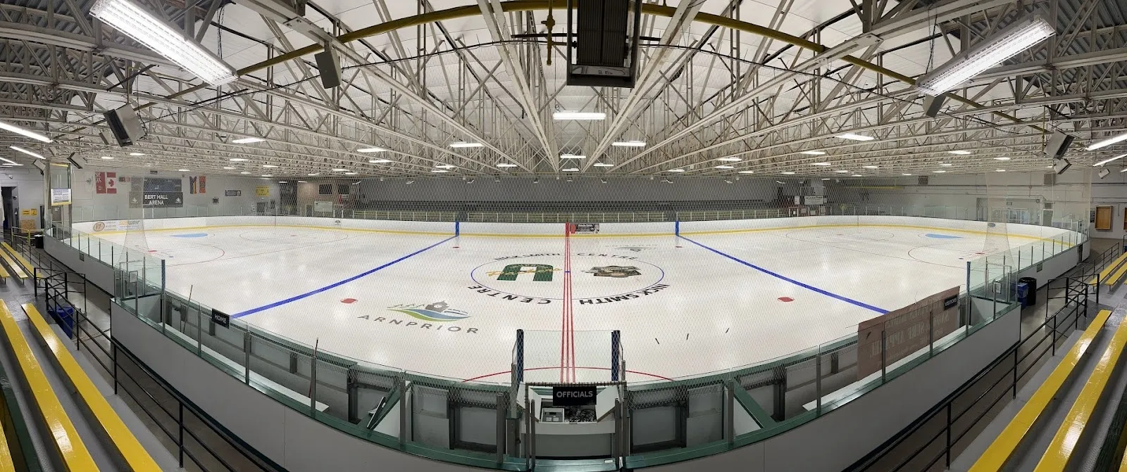 A wide panorama of the empty sheet through curved glass, blue lines and a centre circle under a steel roof with yellow edge boards. — Nick Smith Centre