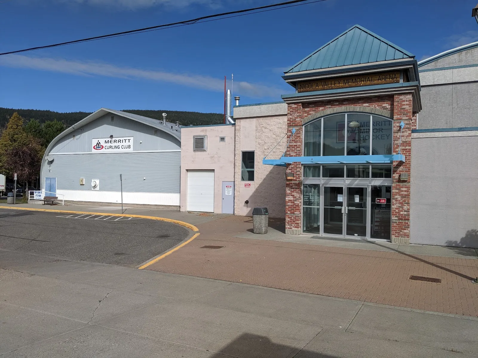 The red-brick entrance tower with a teal pyramid roof and sky-blue awning at the Nicola Valley Memorial Arena, with the grey curved roof of the Merritt Curling Club beside it and a wooded hillside behind. — Nicola Valley Memorial Arena