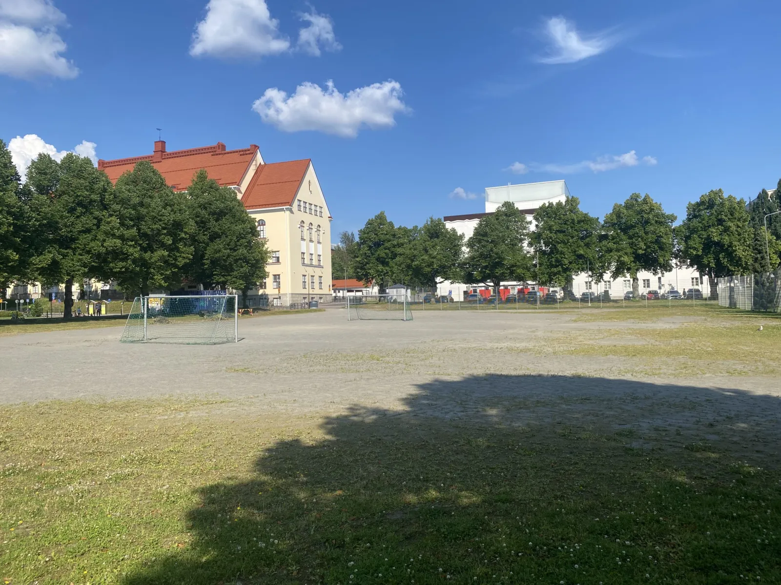 The gravel field beside the Niirala school in Kuopio, which serves as a skating field in winter, seen in summer with the yellow school building and trees behind. — Niiralan luistelukenttä