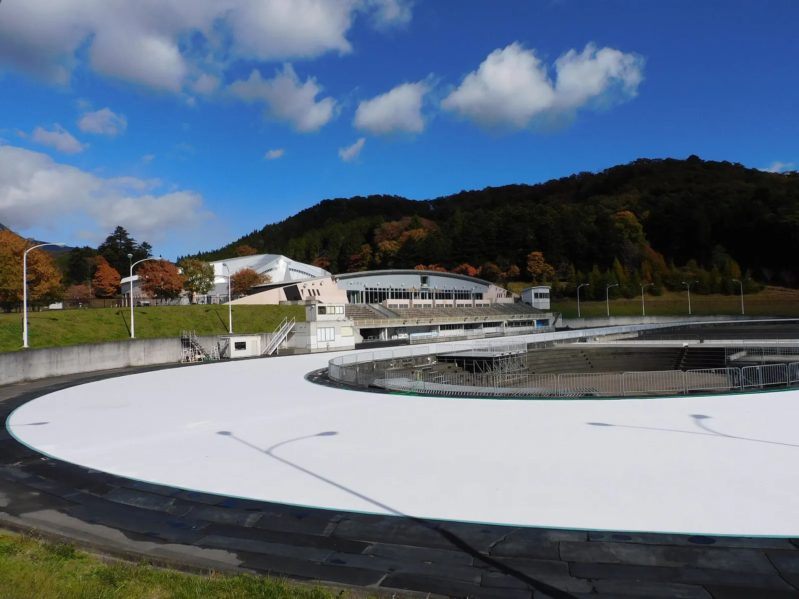 The white oval of the Nikko Kirifuri Skate Center speed-skating rink in autumn, with the grandstand buildings and wooded hills behind. — Nikko Kirifuri Skate Center (outdoor speed rink)