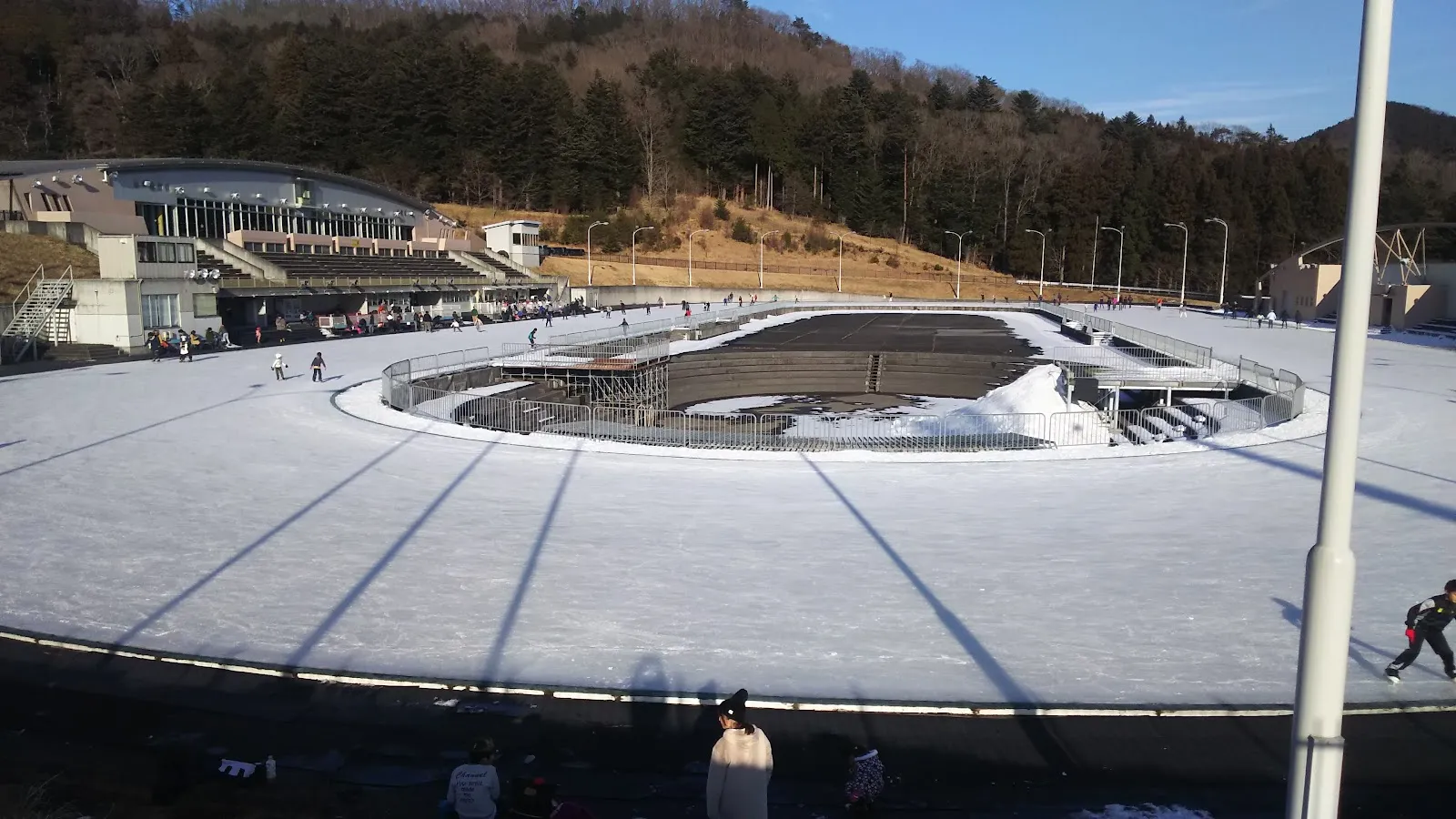 Skaters far out on the frozen outdoor oval at the Nikko Kirifuri Skate Center, with the stands and wooded hills beyond. — Nikko Kirifuri Skate Center (outdoor speed rink)