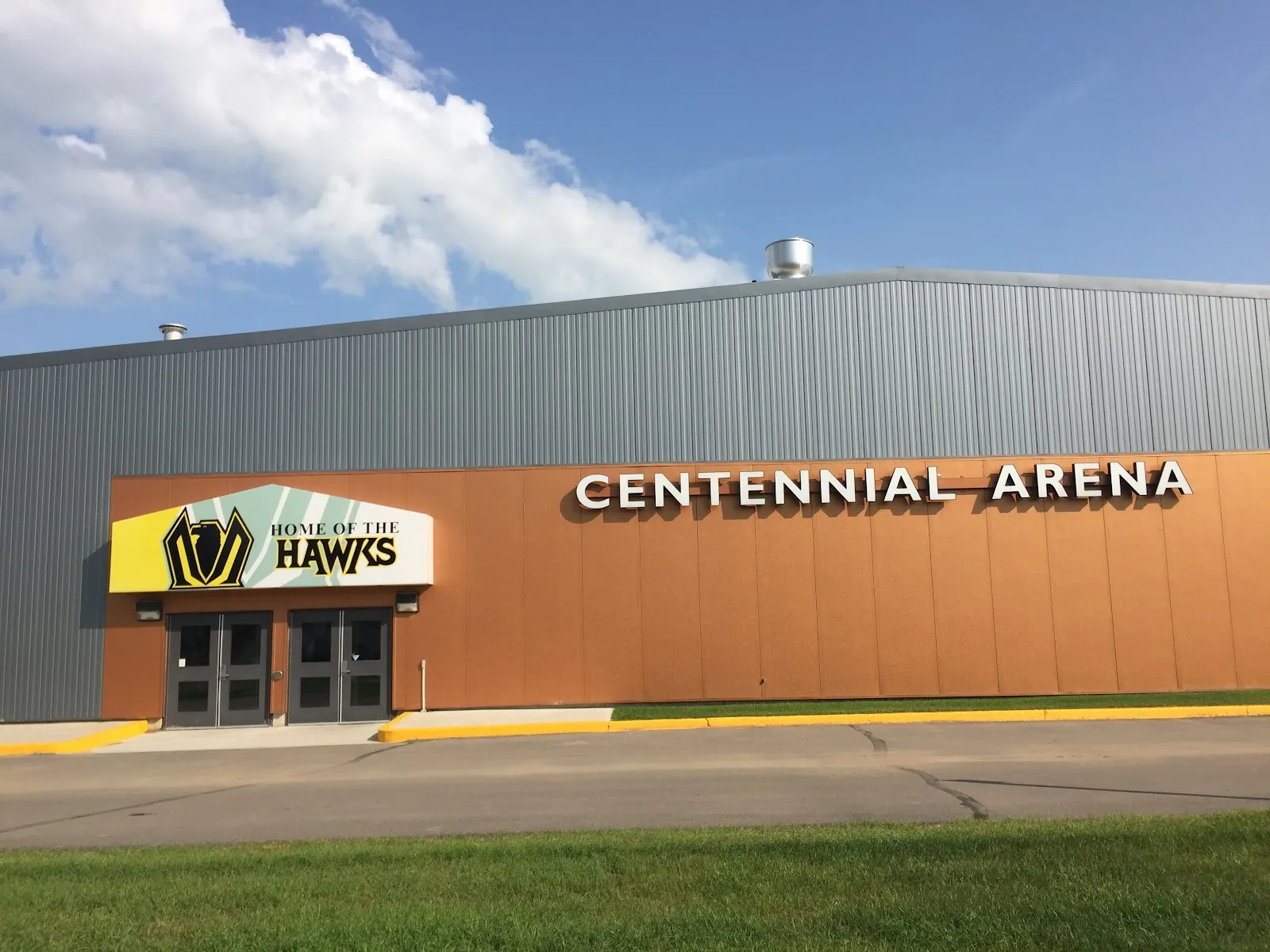 El muro de la entrada de la Centennial Arena en verano: una fachada naranja con las letras de Centennial Arena y un letrero de Home of the Hawks sobre las puertas, bajo un techo metálico gris — Nipawin Centennial Arena