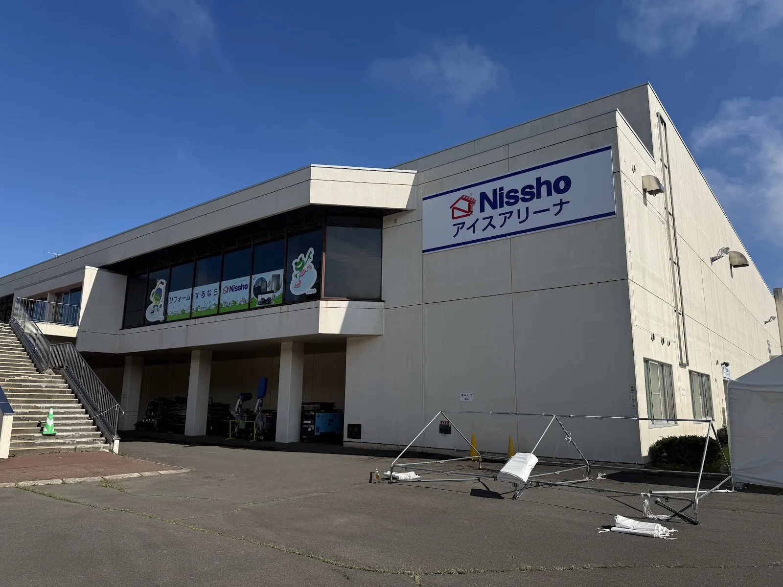 The pale concrete Nissho Ice Arena building with its blue Nissho sign on the end wall, seen from the car park under a blue sky. — Nissho Ice Arena (Kushiro Harutori Ice Arena)