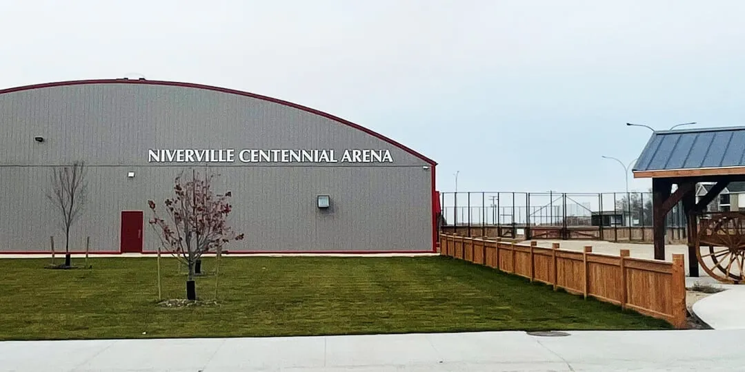 The long side of the Niverville Centennial Arena with its name across the metal wall, a timber fence and a covered pavilion beside it. — Niverville Centennial Arena