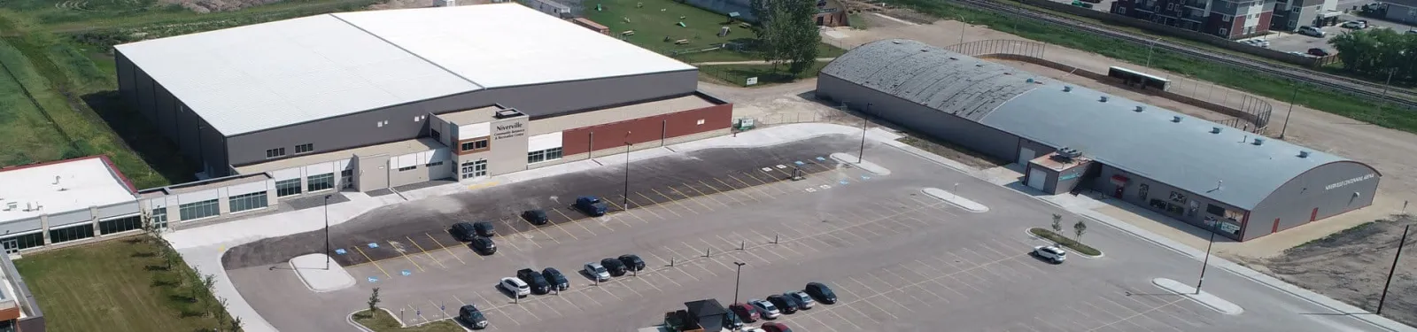 Aerial view of the CRRC's large white-roofed building and parking lot, with the older curved-roof Centennial Arena beside it. — Niverville CRRC