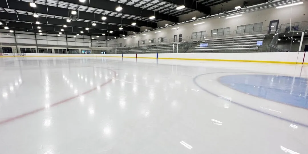 The empty CRRC ice with a blue centre-ice circle, grey stands along one side and white roof trusses above. — Niverville CRRC