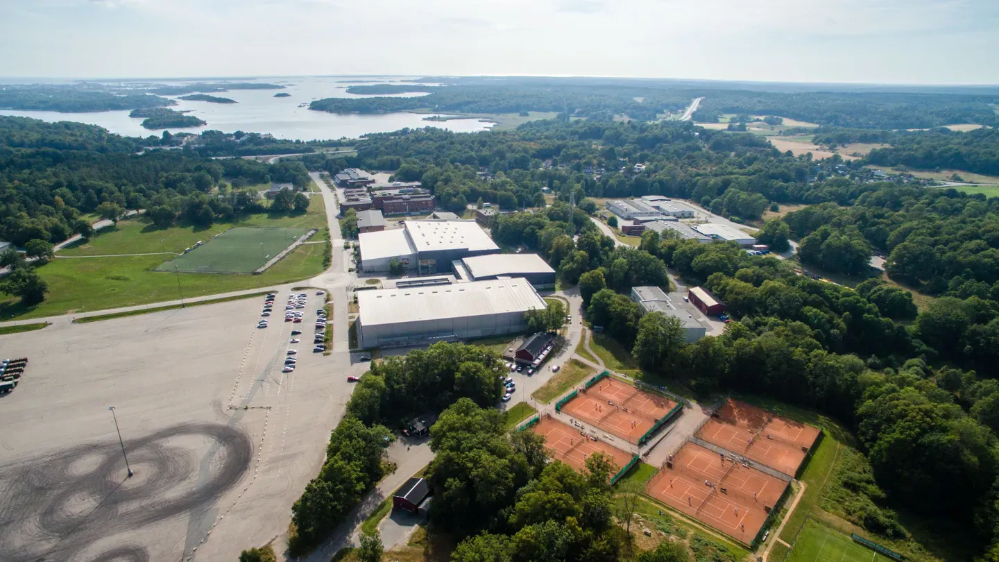 Aerial view of the Rosenholm sports area outside Karlskrona, with the NKT Arena halls at the centre, the tennis courts below them and the archipelago behind — NKT Arena