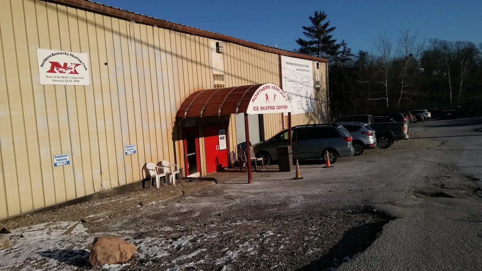 Edificio con revestimiento metálico color arena con una entrada de puertas rojas bajo un toldo curvo que dice NORTHERN KENTUCKY ICE SKATING CENTER, con una manta de NKSC en la pared y autos en el estacionamiento de grava — Northern Kentucky Ice Center