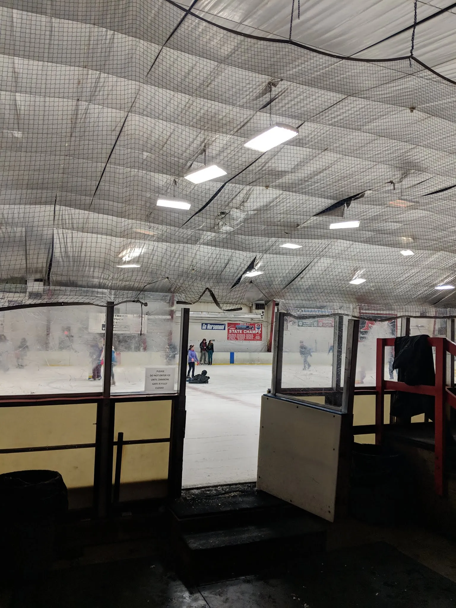 La pista vista a través de la puerta abierta de la banca de castigo, con algunos patinadores en el hielo y una manta de Go Norsemen! STATE CHAMPS en las vallas — Northern Kentucky Ice Center