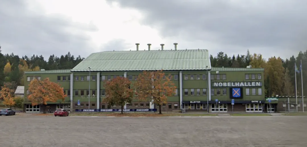 The green Nobelhallen building with its pitched roof and name sign, seen across the car park — Nobelhallen