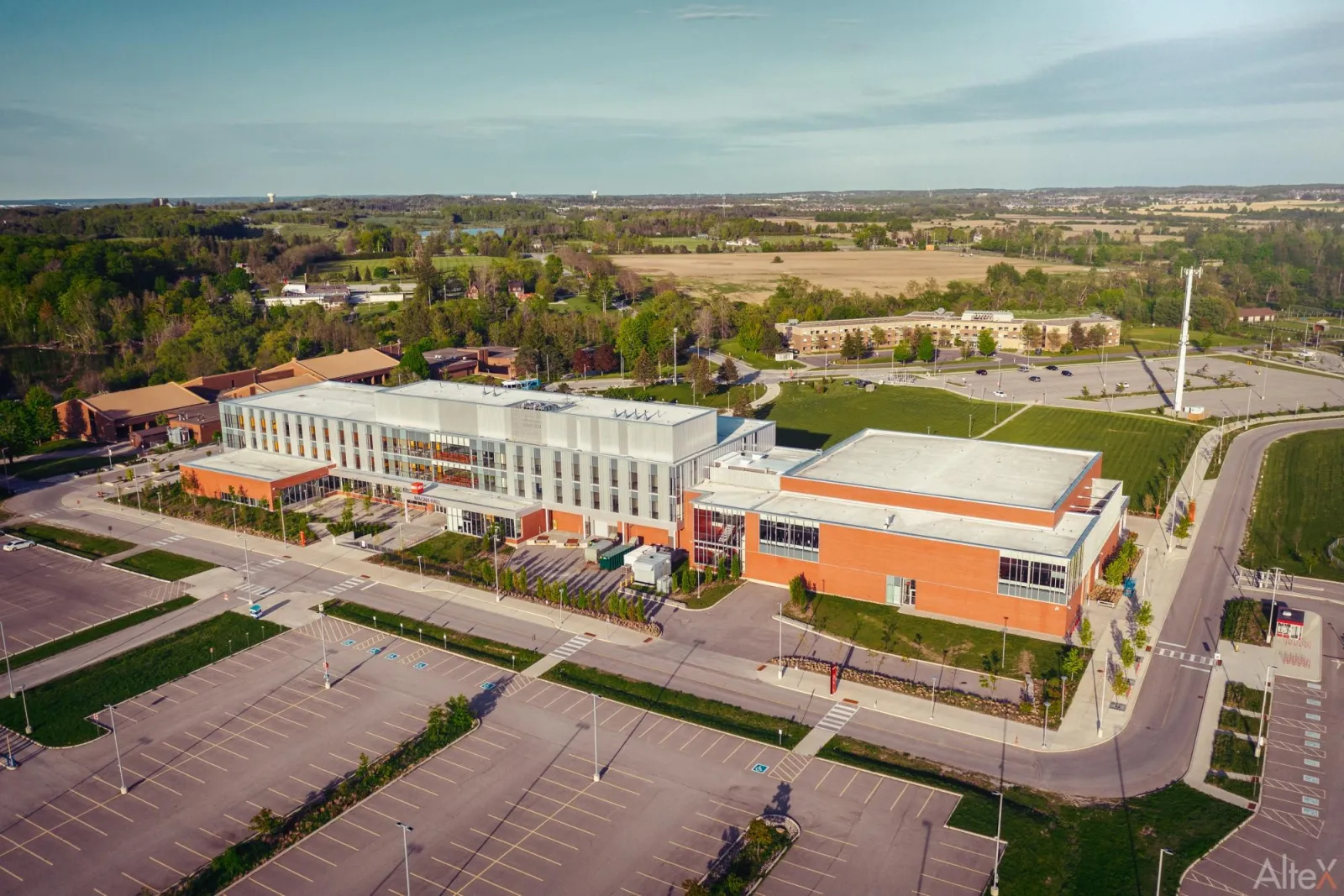 An aerial view of the Dr. William Laceby Nobleton Community Centre and Arena, a long glass-fronted brick complex with an orange-brick arena wing, a car park and sports fields beside it. — Nobleton Community Centre & Arena