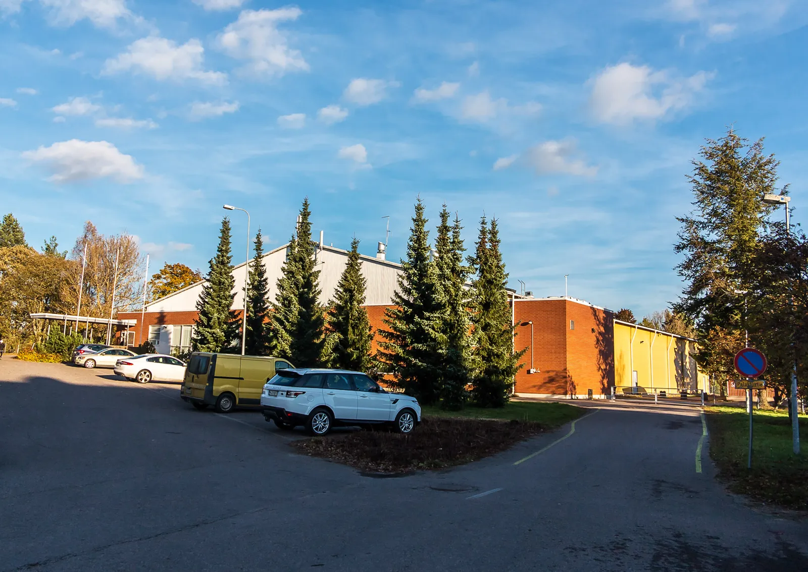 Nokian jäähalli seen across its car park, a low red-brick hall with a pale gable and a yellow wing behind a row of spruces under a blue sky — Nokian jäähalli