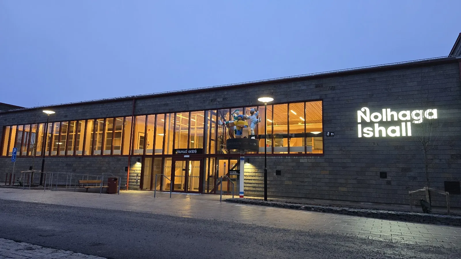 The lit glass entrance of the Nolhaga Ishall in Alingsås at dusk, with the name in illuminated letters on the stone-clad wall — Nolhaga ishall