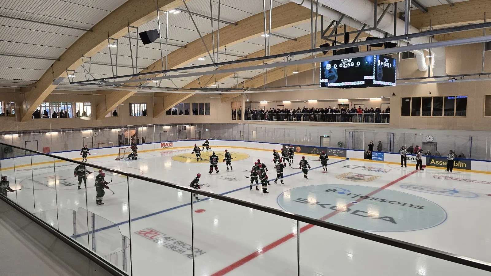 The ice at Nolhaga ishall in Alingsås from the stands, with players at the far end, the scoreboard and the timber-and-glass hall roof. — Nolhaga ishall