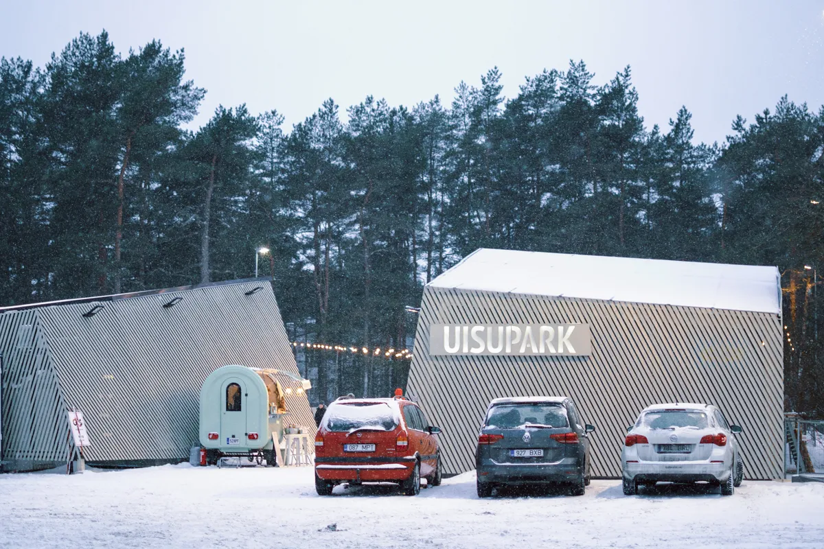 Nõmme välisväljaku teenindushütid mändide vahel, hallil kabiinil UISUPARK-i tähed ja ees lumes seisvad autod. – Nõmme uisupark