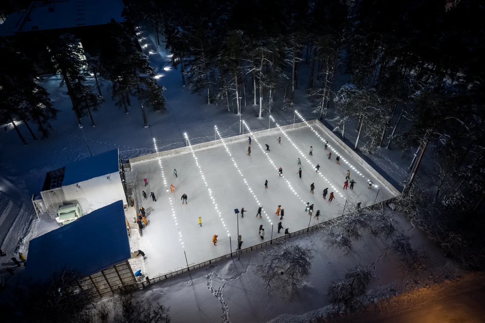 Nõmme välisväljak ülalt öösel, valgustatud jääristkülik lumiste mändide vahel sinise teenindushüti kõrval. – Nõmme uisupark