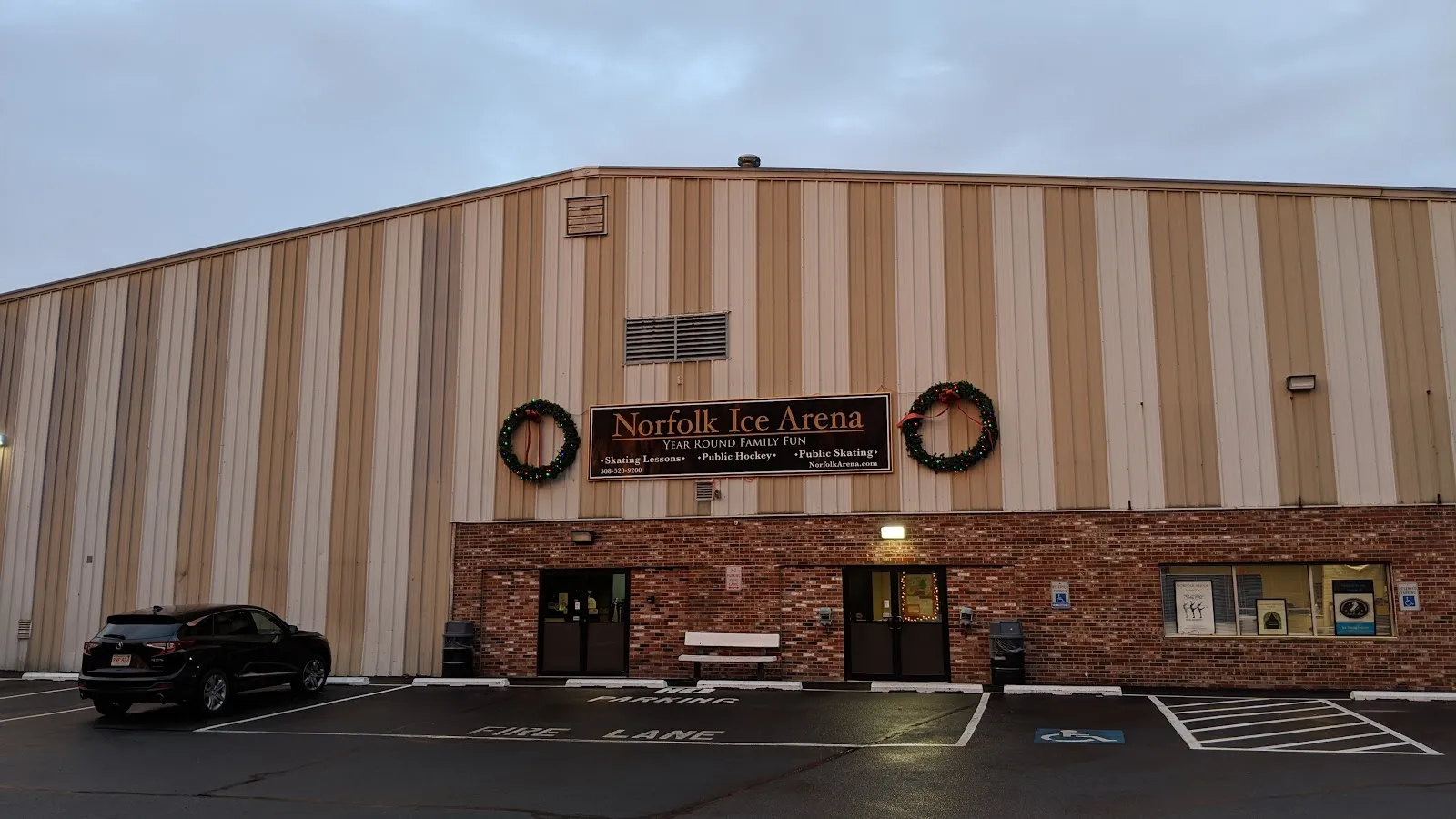The striped metal arena building with a brick base and a "Norfolk Ice Arena" sign between two wreaths over the entrance, with the parking lot in front. — Norfolk Ice Arena