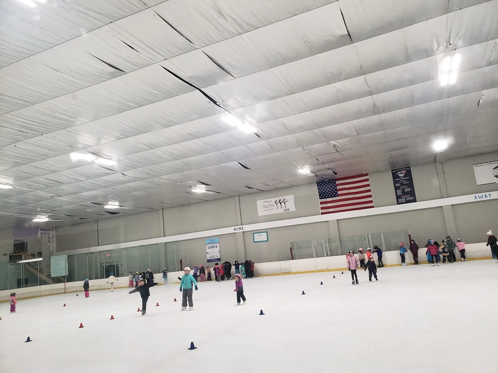 A learn-to-skate session across a wide sheet, skaters spread among cones under a ceiling with lights and an American flag on the far wall. — Norfolk Ice Arena