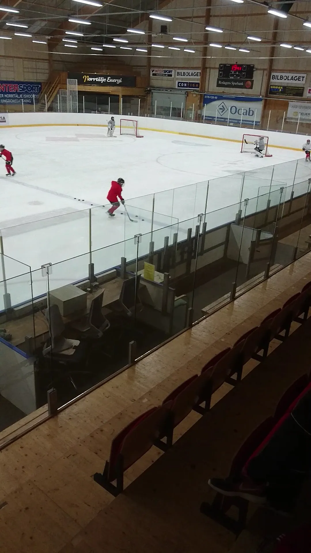 The ice at Norrtälje Sportcentrum seen through the glass from the stands during a match, the players small and far away — Norrtälje Sportcentrum