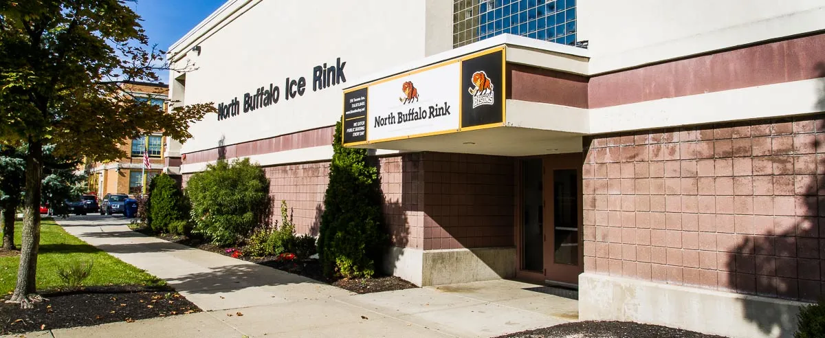 Front entrance of the building, white and pink-brick facade with a large "North Buffalo Ice Rink" wall sign and a lit "North Buffalo Rink" awning sign with the Bisons logo over the door. — North Buffalo Ice Rink