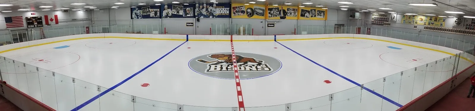 A wide flat view of the sheet with the Buffalo-style centre logo and blue lines, banners hung along the far wall under a low ceiling. — North Buffalo Ice Rink