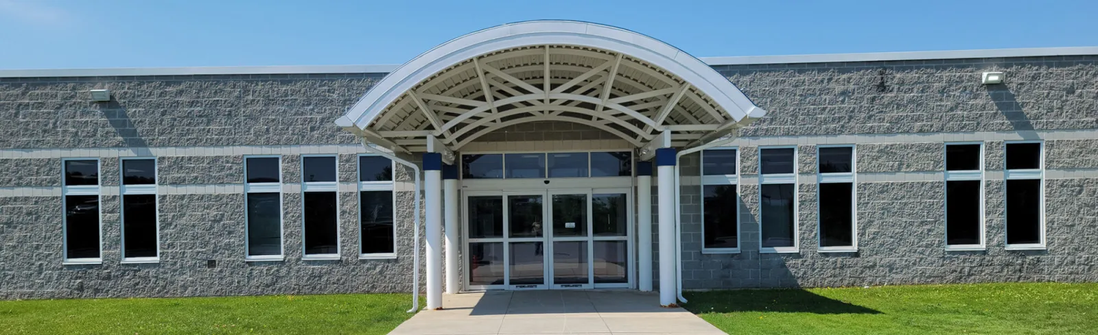 Front entrance of the gray block building, curved white canopy over glass double doors, clear sky — North Central Recreation Center