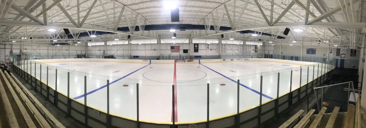 Wide panorama of the full, empty ice sheet, with wooden bleachers along one side and an American flag on the far wall — North Central Recreation Center