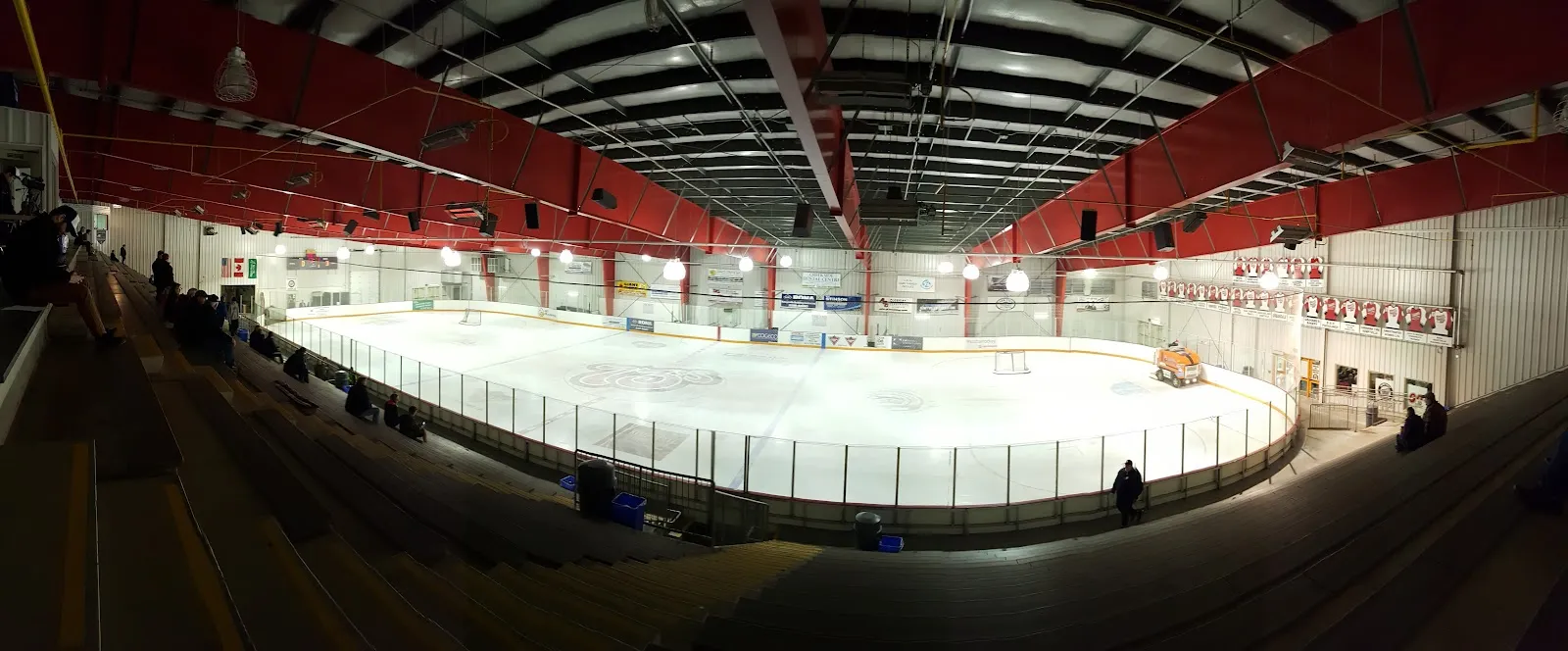A wide panoramic view across the ice sheet from the stands, with red steel roof trusses overhead, the boards below and the ice resurfacer parked at the far end. — North Grenville Municipal Centre
