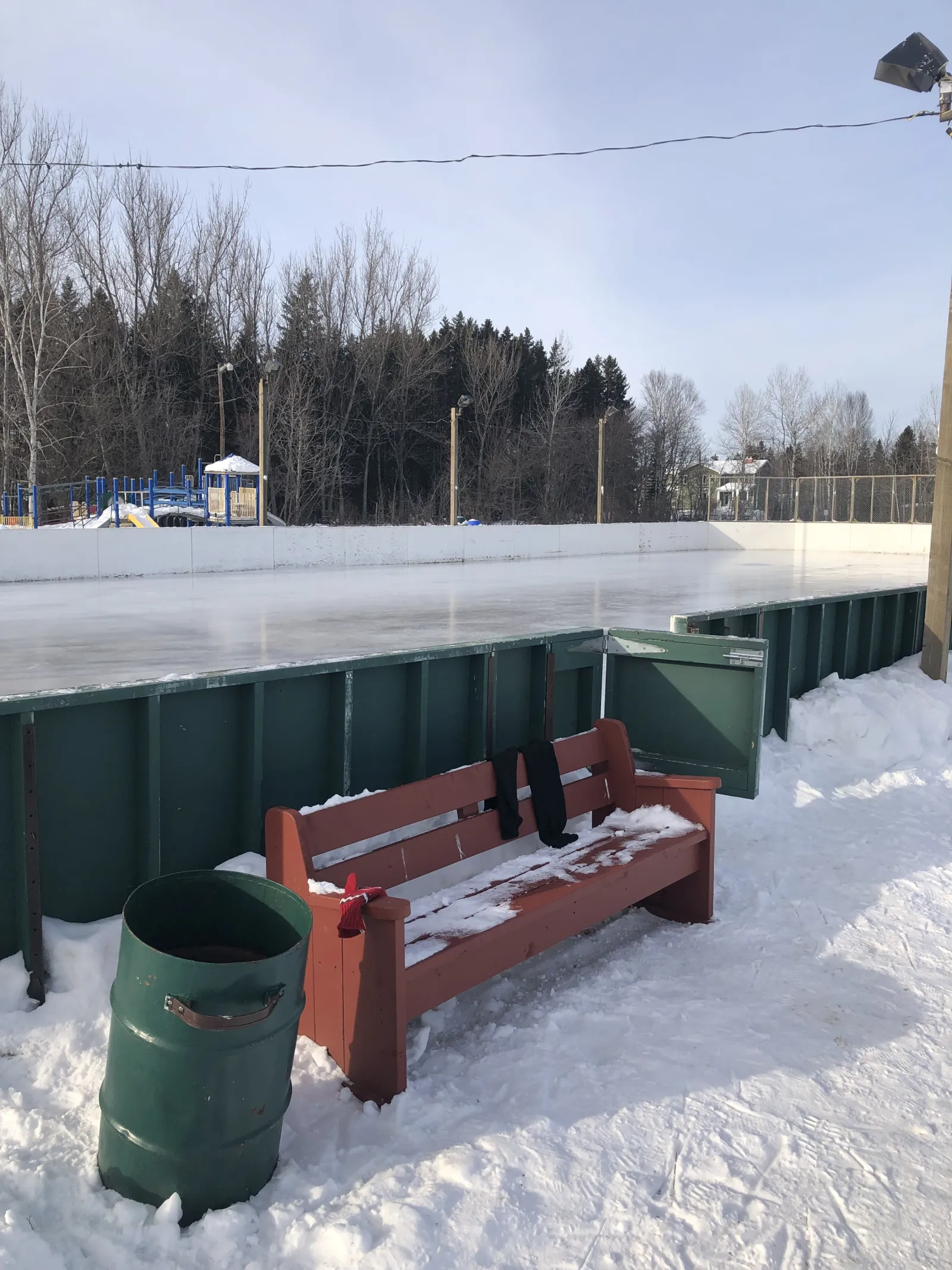 A bench and bin beside the North Neebing Park rink, with the play area beyond — North Neebing Park Outdoor Rink