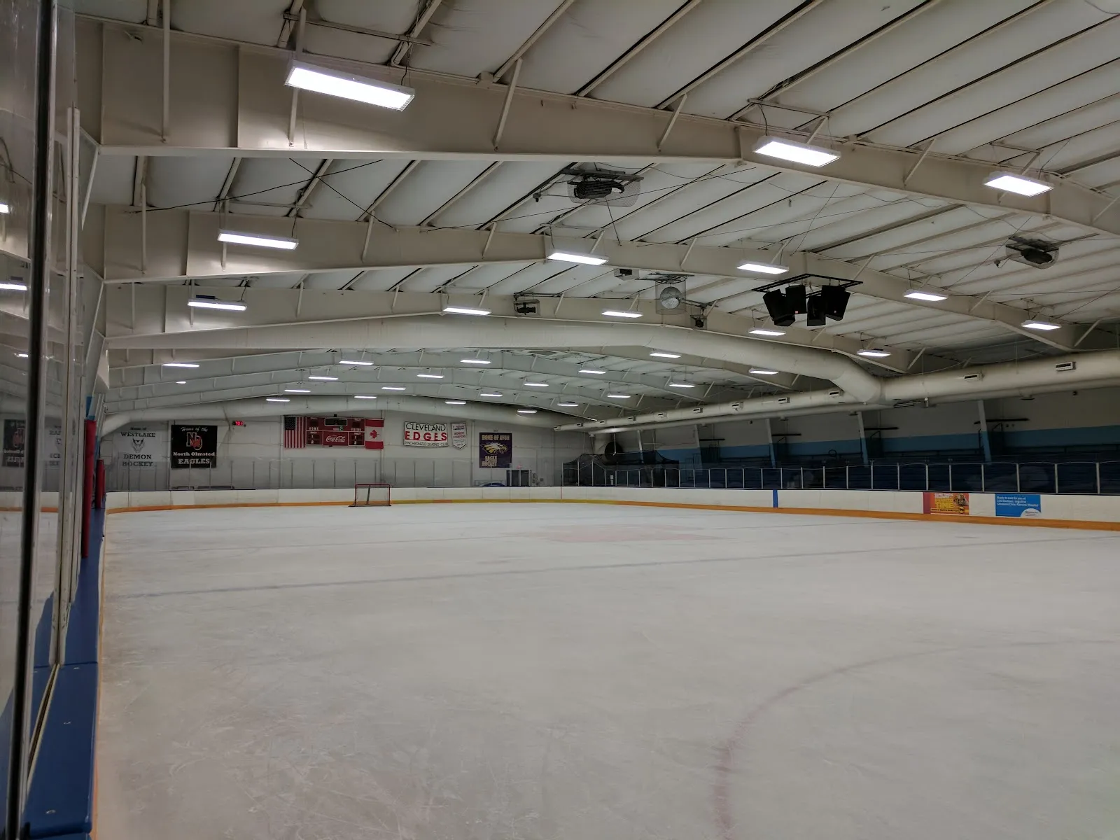 A wide empty sheet of ice under a ceiling of ductwork and lights, blue boards and glass along the left and a flag-lined wall at the far end. — North Olmsted Recreation Center