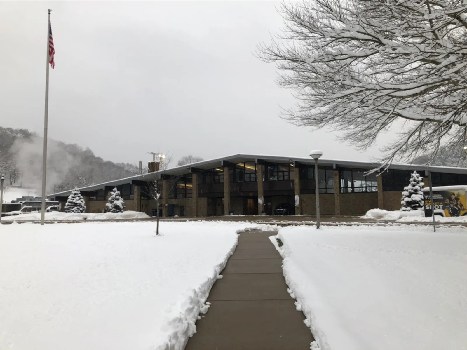 El edificio bajo de madera y vidrio tipo cabaña de North Park Ice Rink bajo la nieve, visto a lo largo del camino despejado desde el asta de bandera — North Park Ice Rink