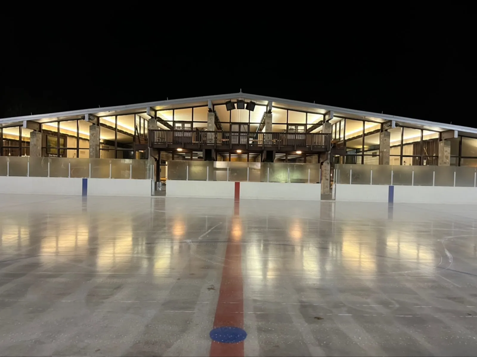 El hielo exterior vacío de North Park Ice Rink de noche, iluminado desde la cabaña con su balcón y su muro de vidrio detrás de las vallas — North Park Ice Rink