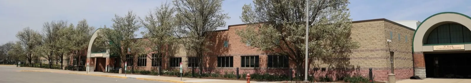Wide street-level panorama of the Northbrook Sports Center: a long tan-brick building with an arched glass entrance at each end, trees along the front lawn. — Northbrook Sports Center