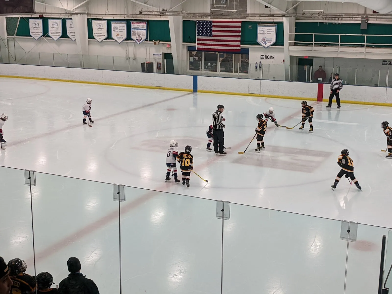 A game seen through the glass, players spread across a wide sheet beneath a large American flag and club banners on the far wall. — Northbrook Sports Center