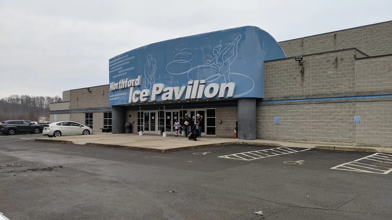 The curved blue front of the Northford Ice Pavilion with its name across the roof edge, a flat grey wall beside it and an empty car park in front. — Northford Ice Pavilion