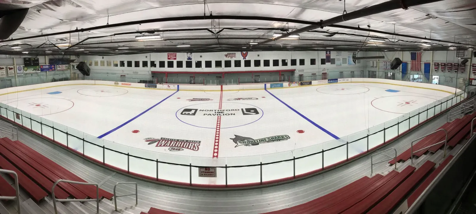 Wide, empty view of the full ice sheet from the stands, with a "Northford Ice Pavilion" logo painted at center ice, team banners, and an American flag. — Northford Ice Pavilion