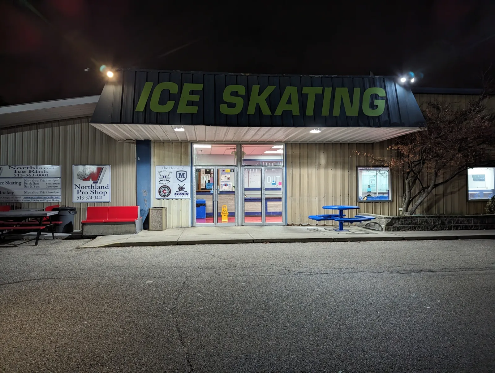 Night exterior of Northland Ice Center's metal-sided building, lit "ICE SKATING" sign above the entrance awning, with "Northland Ice Rink" and "Northland Pro Shop" signage flanking the glass doors and empty picnic tables on the sidewalk. — Northland Ice Center
