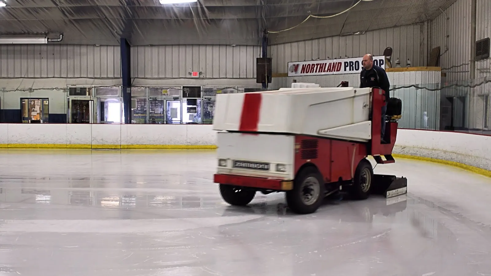 Most of the ice sheet during resurfacing, with a red-and-white Zamboni and its operator on it and a "NORTHLAND PRO SHOP" sign on the far wall; rink boards and metal building interior visible. — Northland Ice Center